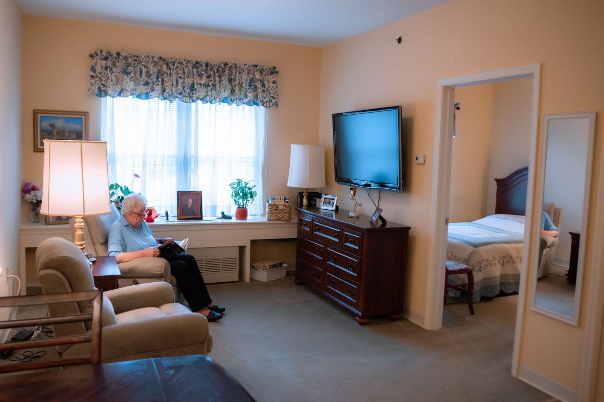 A cozy living room with a person sitting in an armchair, watching TV, and a view into the adjacent bedroom.