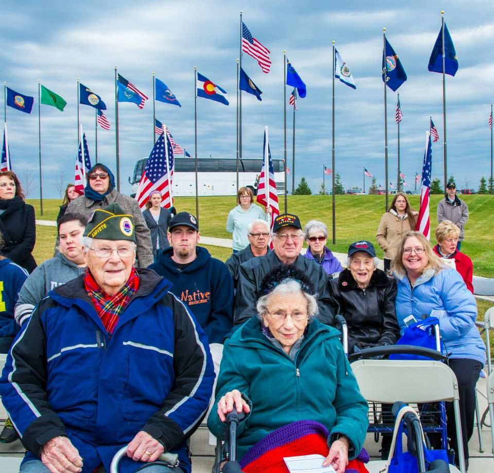 A group of people, including veterans, attending an outdoor ceremony with multiple flags displayed in the background.