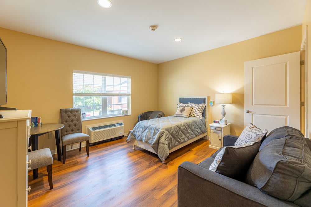 A cozy bedroom with wooden flooring, featuring a bed, a sofa, and a window allowing natural light.