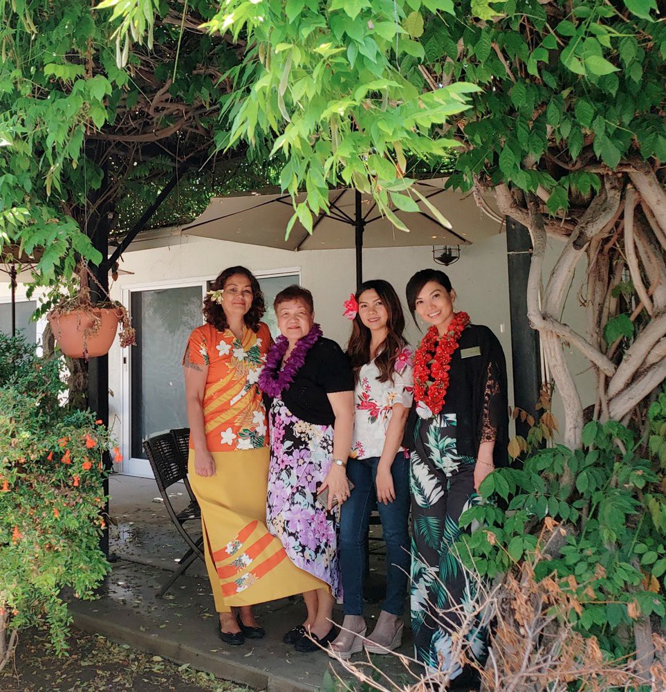 A group of women standing together in a garden, wearing colorful traditional clothing and lei necklaces.