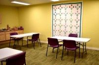 A classroom or meeting room with tables, chairs, and a quilt hanging on a yellow wall.