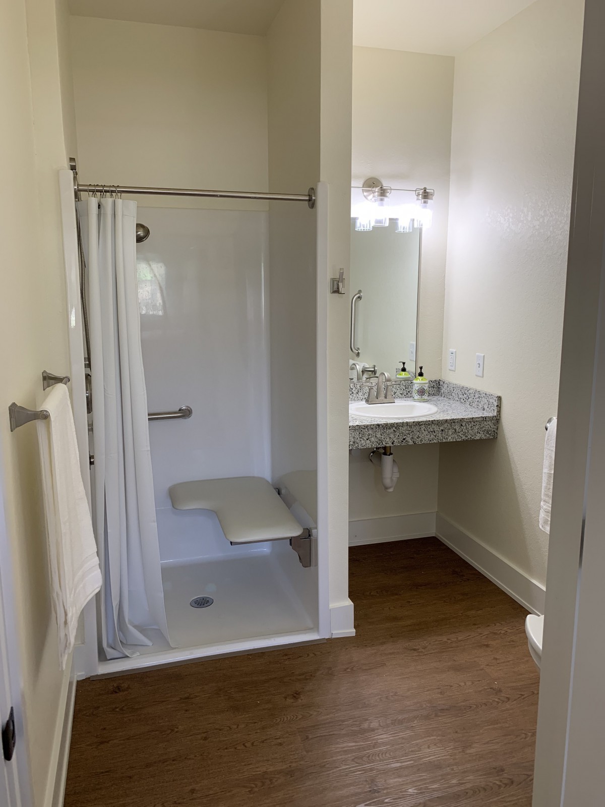 An accessible bathroom with a shower featuring a folding seat and a sink with a mirror.