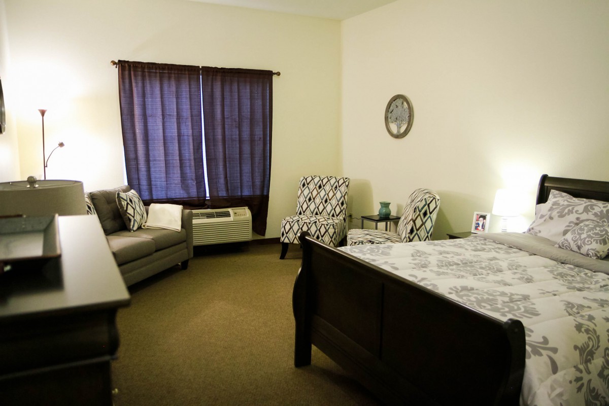 A cozy bedroom with a bed, sofa, and two patterned chairs adjacent to a window with dark curtains.