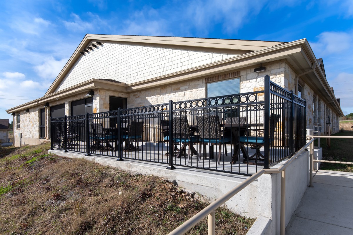 A single-story building with an outdoor patio area featuring black railings and seating, set against a clear blue sky.