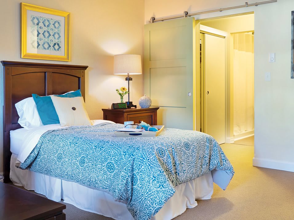 A cozy bedroom with a blue-patterned bedspread, a nightstand with a lamp, and an open sliding door.