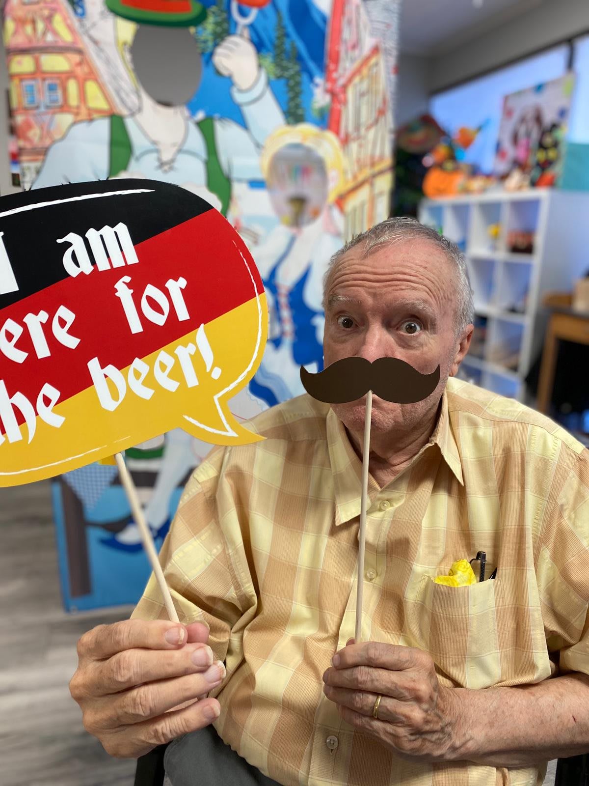 A person playfully holds a sign and mustache prop, celebrating with a humorous 'I am here for the beer!' message.