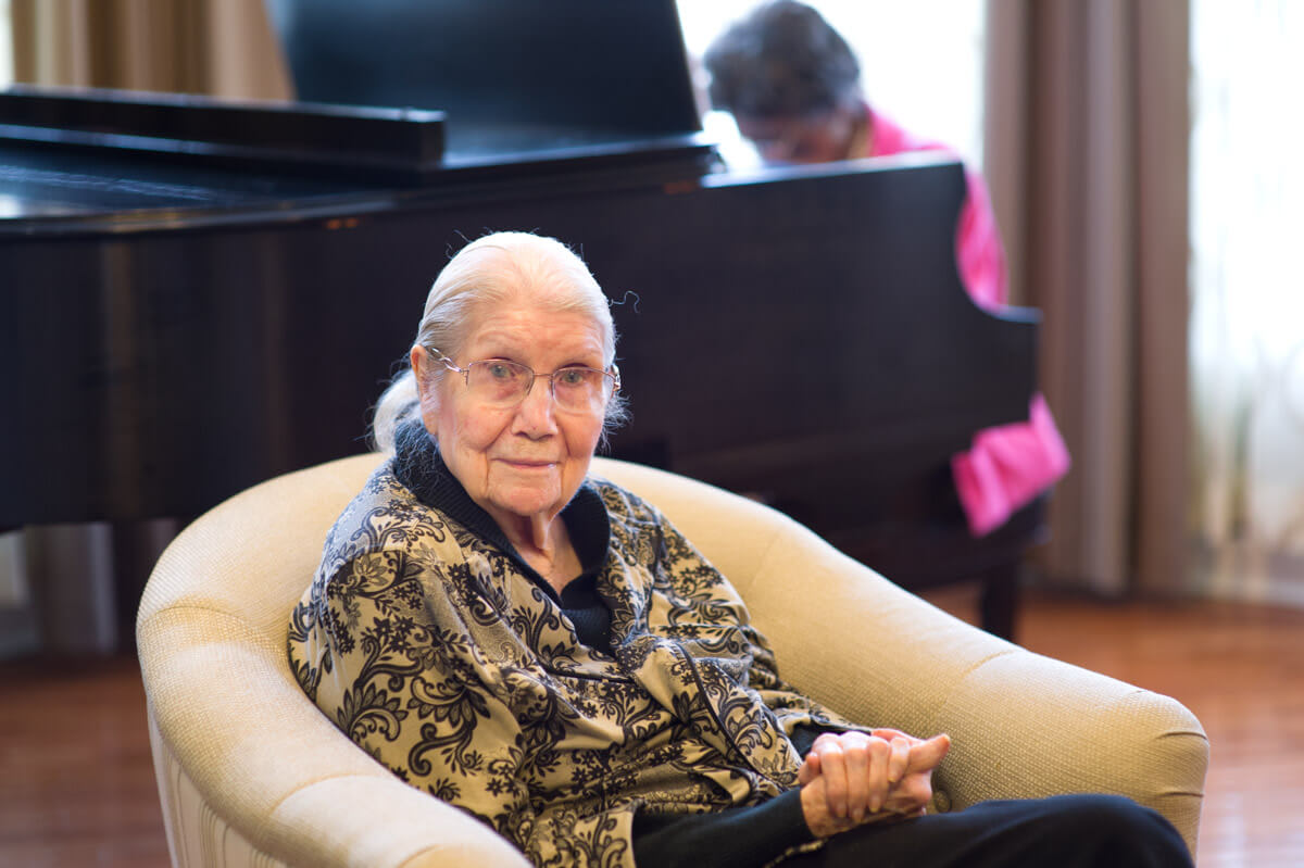 An elderly woman sitting comfortably in a chair with a piano in the background.