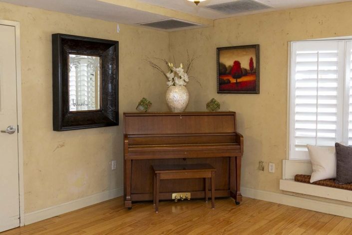 A cozy room featuring an upright piano with a bench, a large vase with flowers on top, and decorative paintings and mirror on the walls.