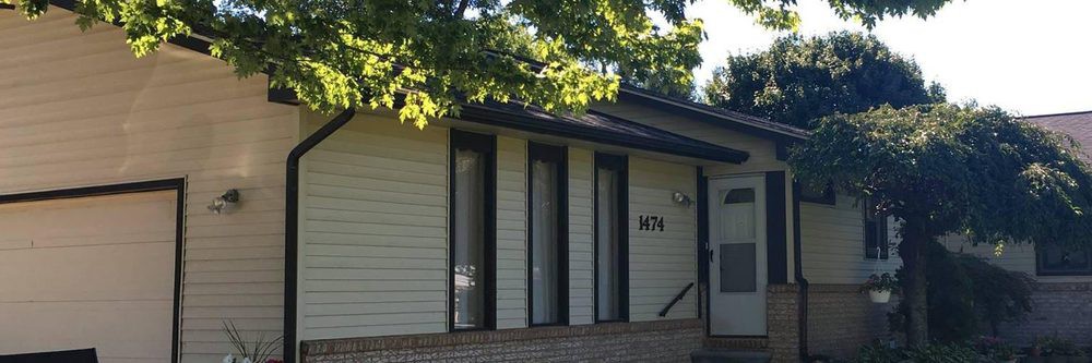 A residential house with a light exterior and narrow windows, partially shaded by trees.