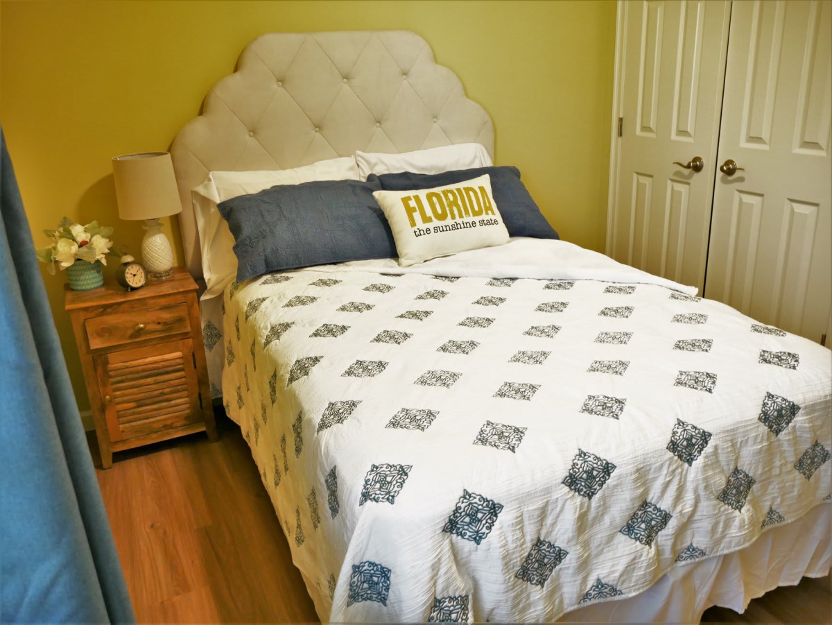 A cozy bedroom with a neatly made bed, featuring a quilt and decorative pillows, next to a wooden nightstand with a lamp.