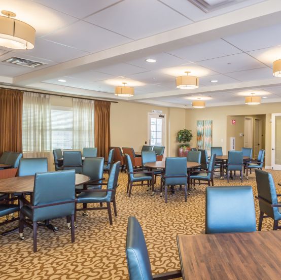 A spacious dining room with multiple tables and chairs, featuring carpeted flooring and modern lighting.