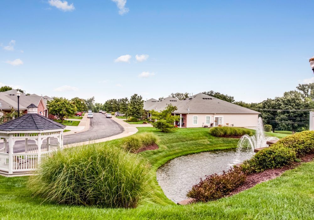 A peaceful suburban neighborhood with well-maintained houses, lush greenery, and decorative fountains along the road.