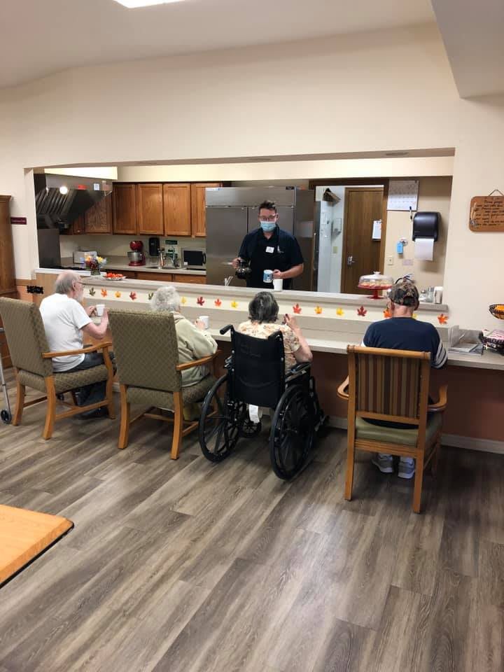 Residents gather in a care facility dining area, interacting with staff.