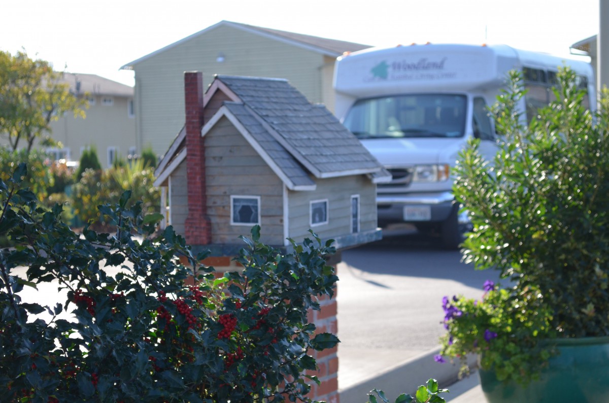 A miniature house mailbox sits beside a driveway with an RV parked in the background.