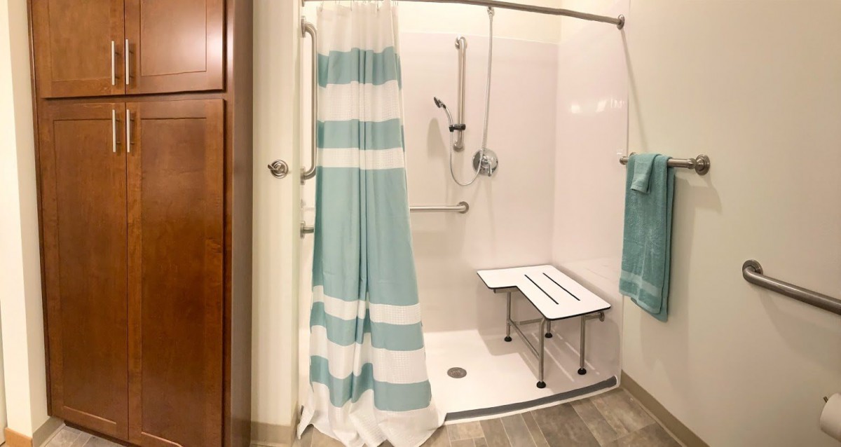 An accessible bathroom shower with a blue and white striped curtain, equipped with a shower bench, handheld showerhead, and safety grab bar.