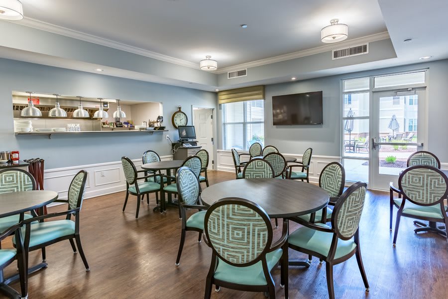 A well-lit dining area featuring several round tables and cushioned chairs, with a television mounted on the wall and large windows providing natural light.