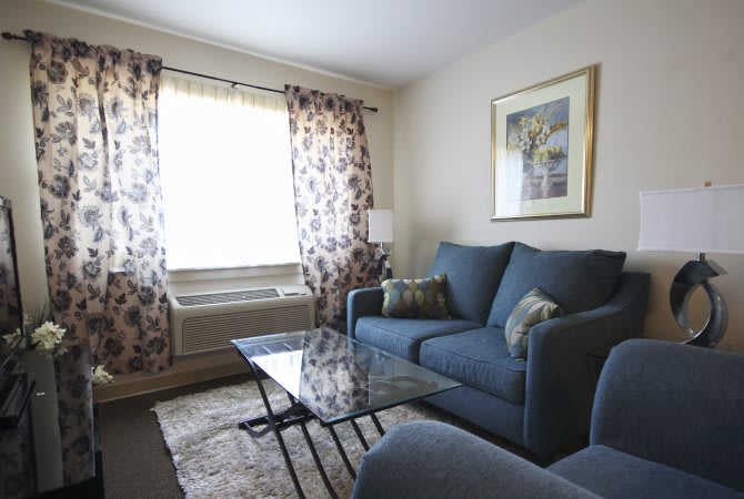 A cozy living room with a blue sofa set, glass coffee table, and patterned curtains.