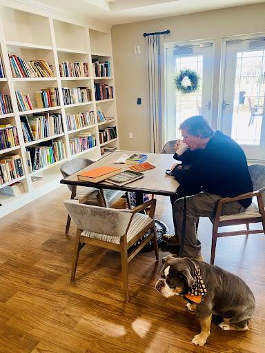 A man sits thoughtfully at a table surrounded by books, with a dog resting nearby.