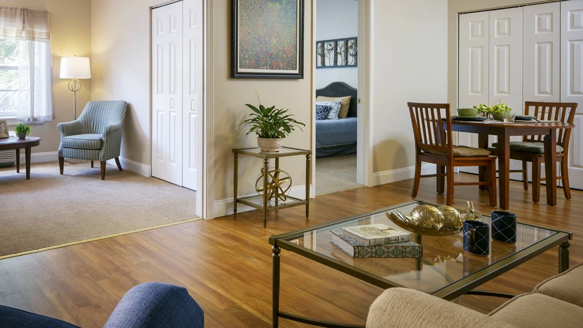 A cozy living room with wooden floors, featuring a coffee table, plant, and artwork on the wall.