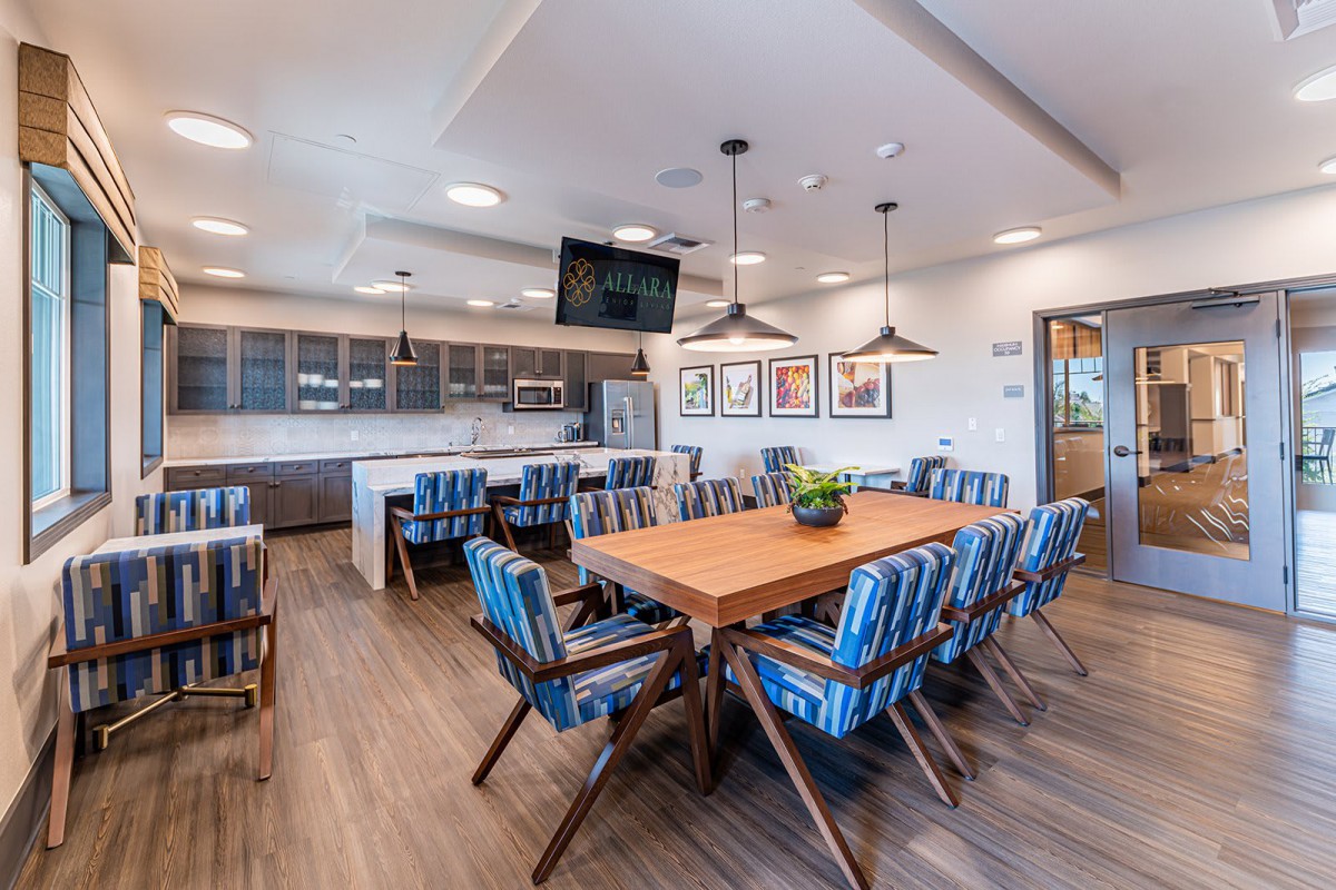A modern dining and kitchen area featuring a large wooden table surrounded by blue-striped chairs, with pendant lighting overhead.
