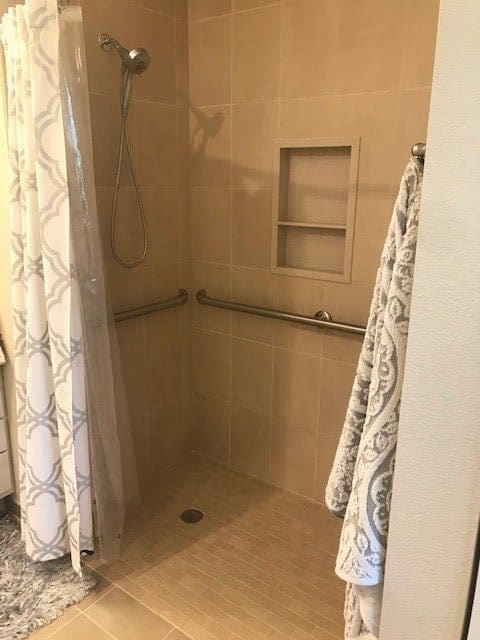 A walk-in shower with a tiled interior and grab bars, featuring a patterned curtain and recessed shelf.