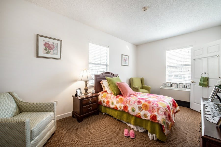 A cozy bedroom with a floral-patterned bedspread, a nightstand with a lamp, and two windows providing natural light.