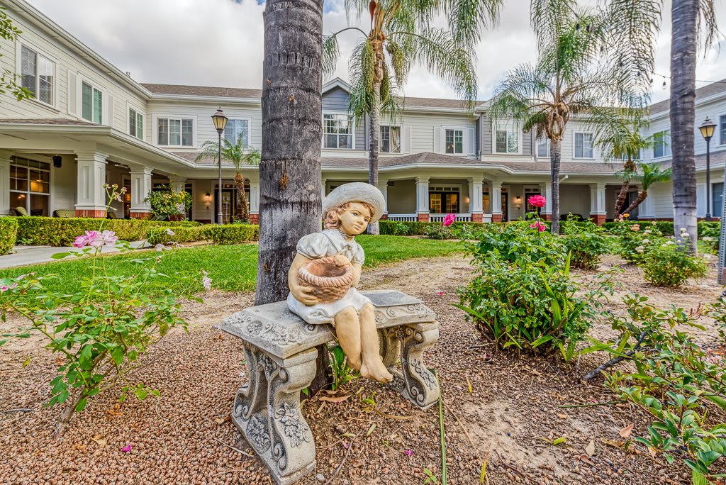 A garden setting featuring a sculpture of a girl sitting on a decorative bench, surrounded by lush greenery and palm trees in front of a large building.