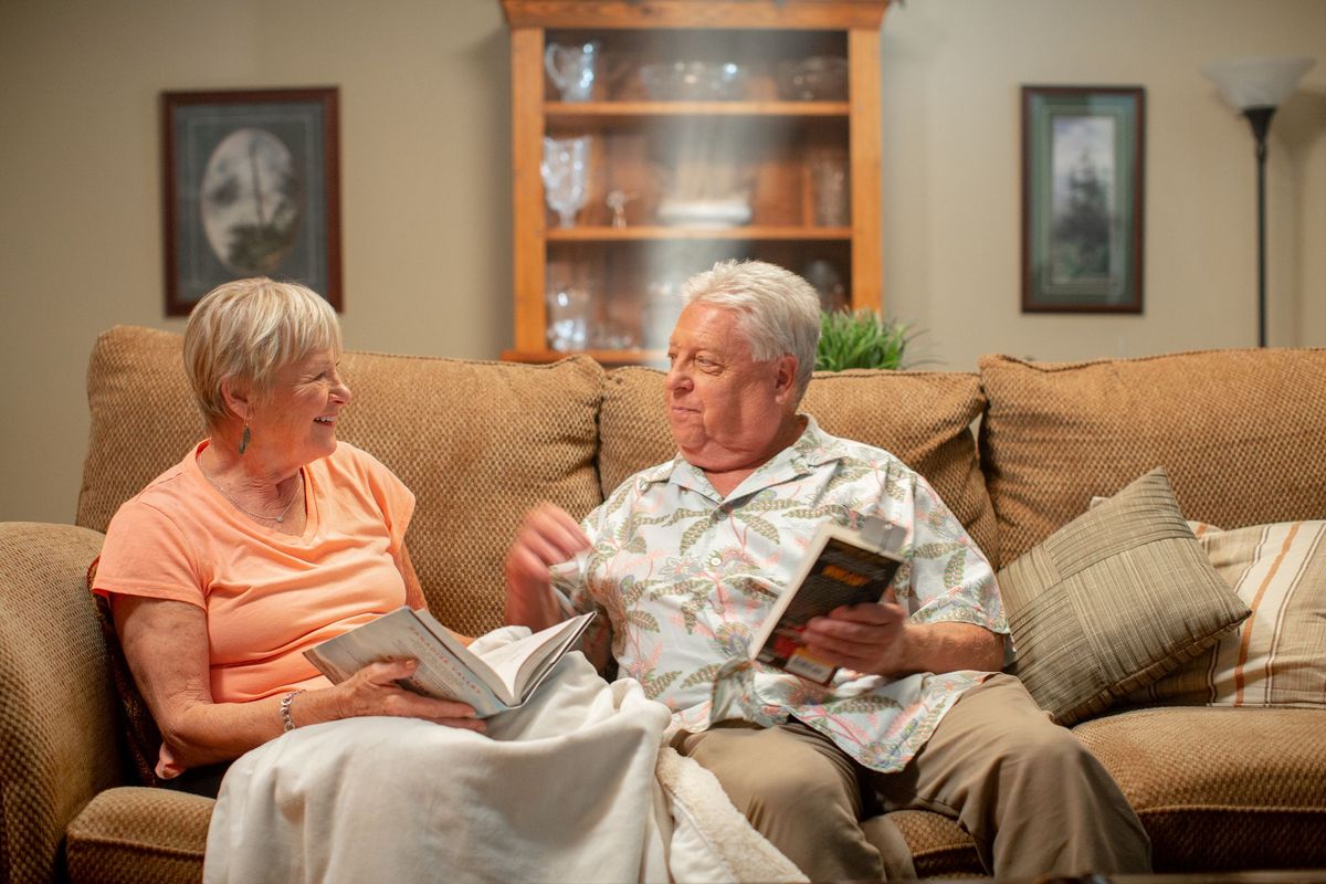 An elderly couple sits on a couch in their living room, engaged in conversation while holding books.