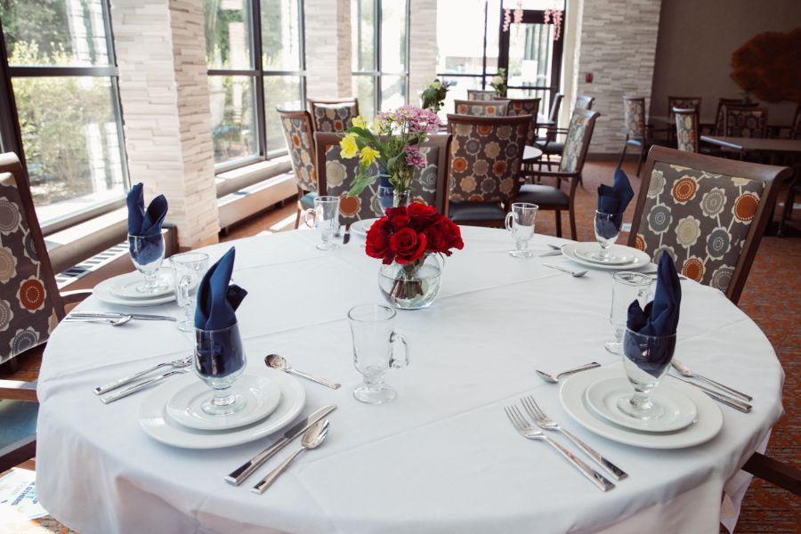 An elegantly set dining table with a white tablecloth, adorned with red and yellow flowers, blue napkins, fine china, and polished silverware.