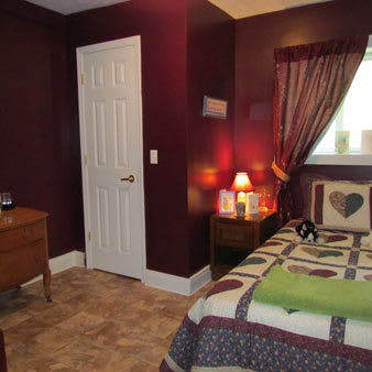 A cozy bedroom with deep red walls, a bed adorned with patterned bedding, soft lighting from a lamp, and curtains framing a window.