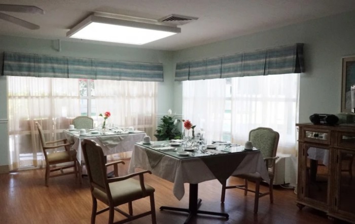 A well-lit dining room with neatly set tables and chairs, adorned with simple decor and surrounded by large windows.