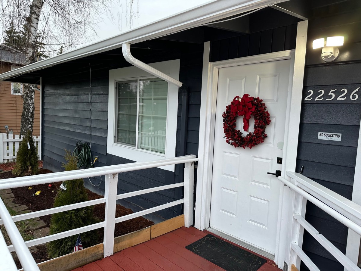 A house with a white door adorned with a red wreath, featuring a window and a porch railing.