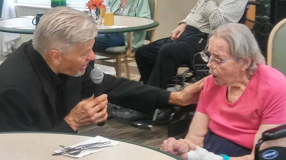 An elderly man holding a microphone engages with a senior woman in a care facility.