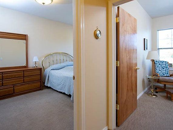 View of a bedroom and hallway through an open door, featuring a bed, dresser with a mirror, and a lamp.