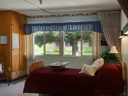 A cozy bedroom featuring a red bedspread, large windows with blue curtains, and warm wooden furniture.