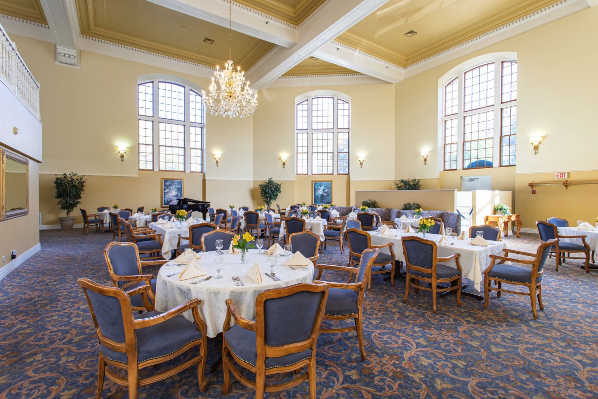 A spacious dining hall with elegantly set tables and comfortable chairs, illuminated by a grand chandelier and large windows.