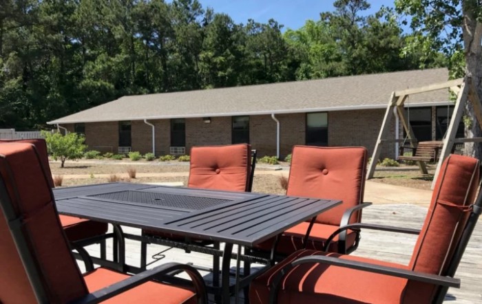 Outdoor patio set with orange cushioned chairs and a metal table, in front of a brick building surrounded by trees.