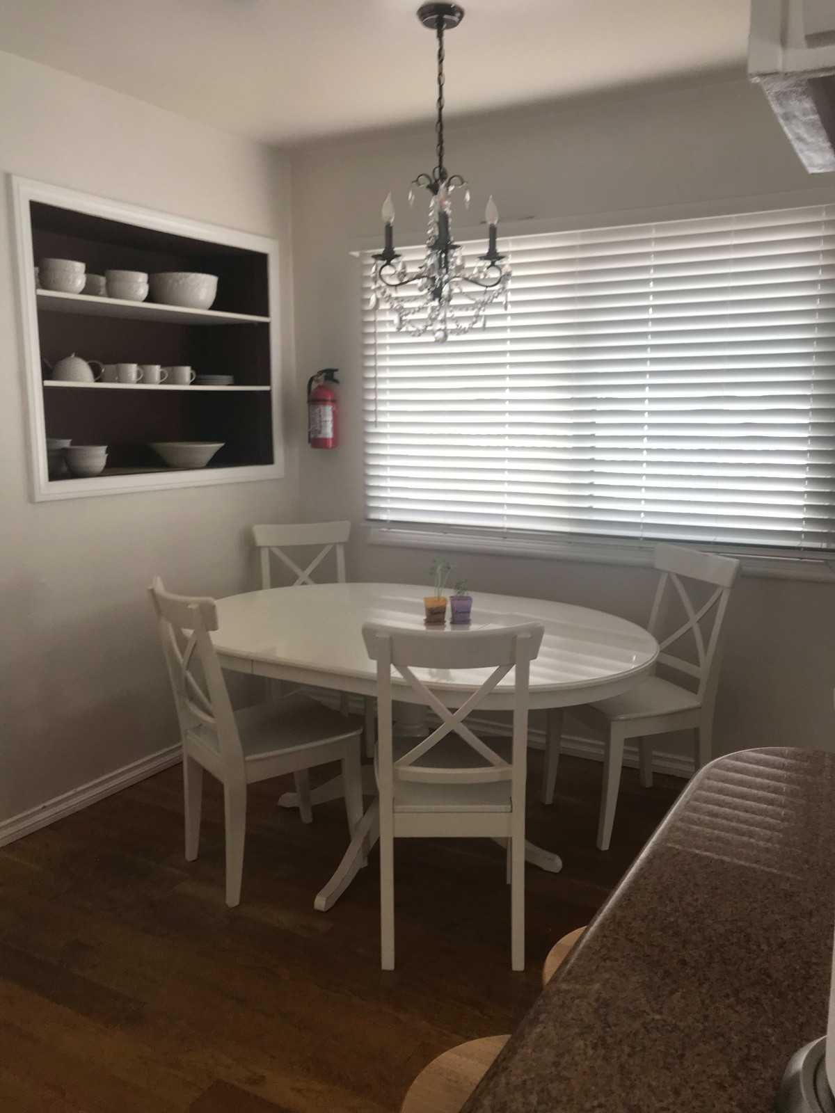 A cozy dining area with a round white table, four matching chairs, and a chandelier overhead. Natural light streams in through the window blinds, highlighting the neatly arranged dishes on open shelves.