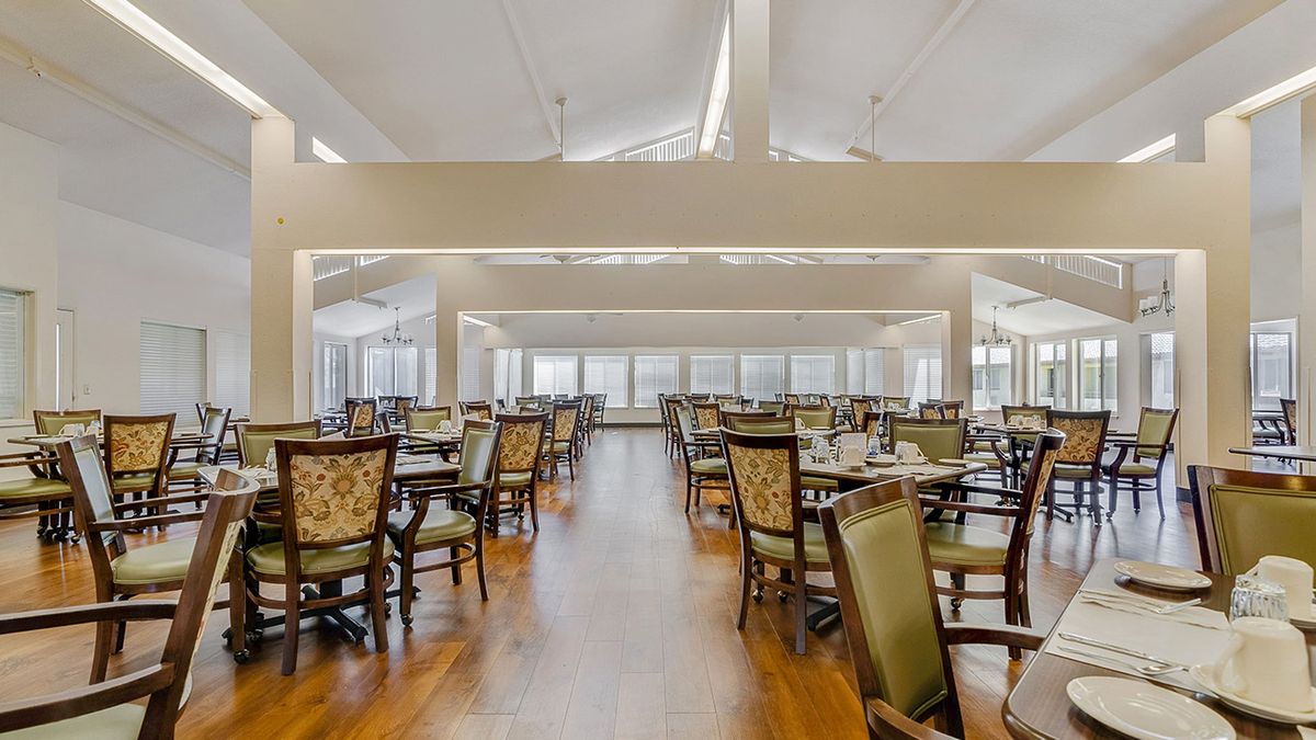 A spacious dining room with neatly arranged tables and chairs, featuring large windows and a hardwood floor.