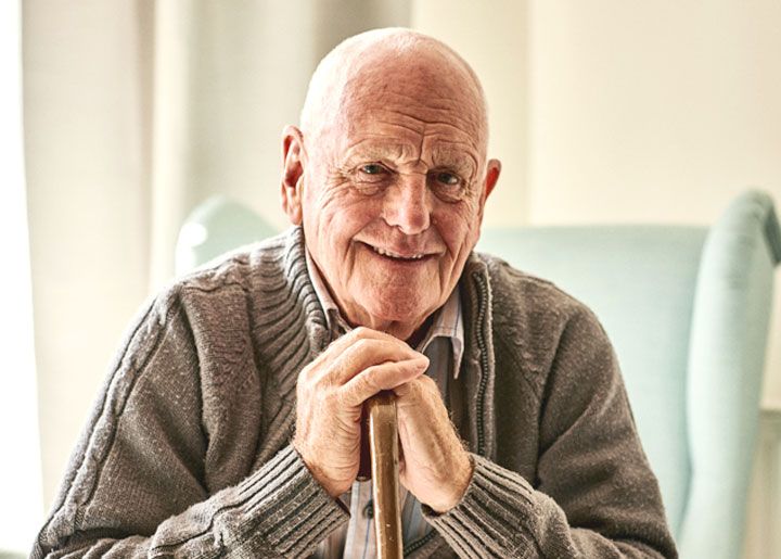 An elderly man smiling warmly while sitting indoors, wearing a grey sweater.