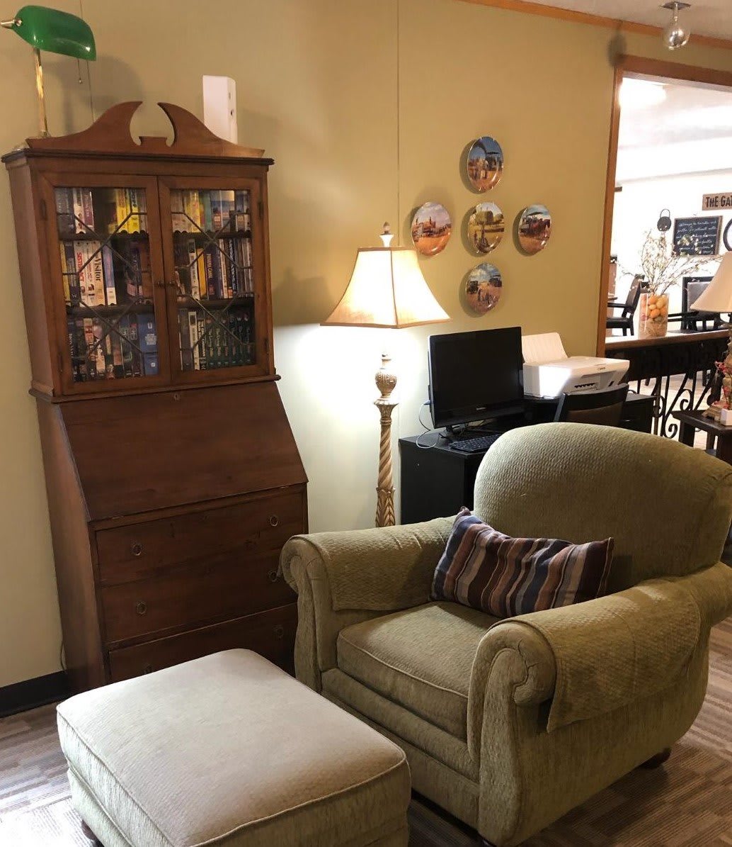 A cozy living room corner with an armchair, ottoman, and a wooden bookshelf, accompanied by a floor lamp and decorative plates on the wall.