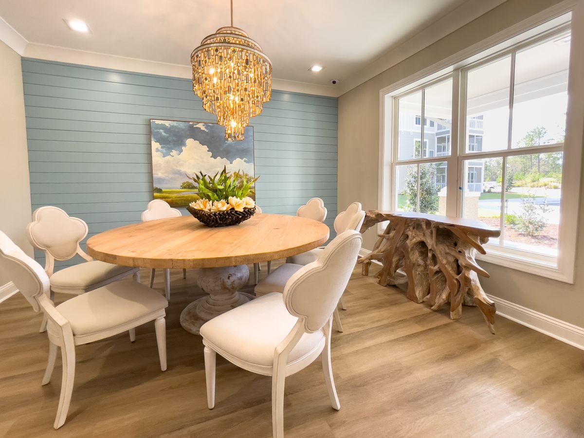 A modern dining room featuring a round wooden table, white chairs, and pendant lighting, complemented by wood flooring and abundant natural light.