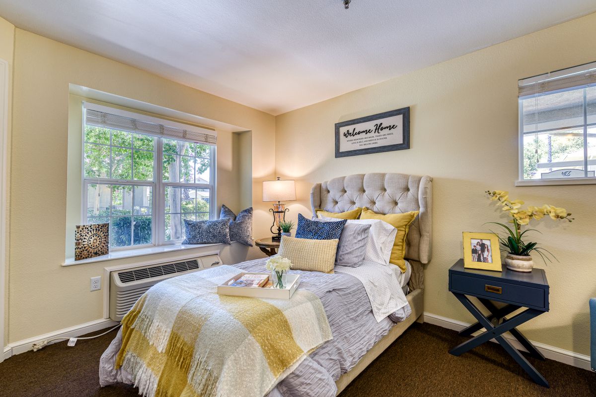 A cozy bedroom with a neatly made bed, featuring pillows and a blanket in yellow and gray tones, illuminated by a bedside lamp next to a large window.
