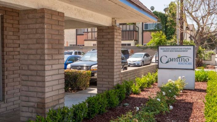 A parking lot with several cars parked beside a brick building, featuring neatly trimmed bushes and a sign.