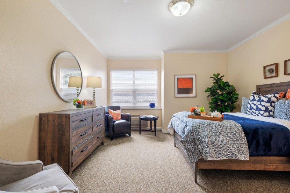 A cozy bedroom with a bed, dresser, and armchair next to a window.