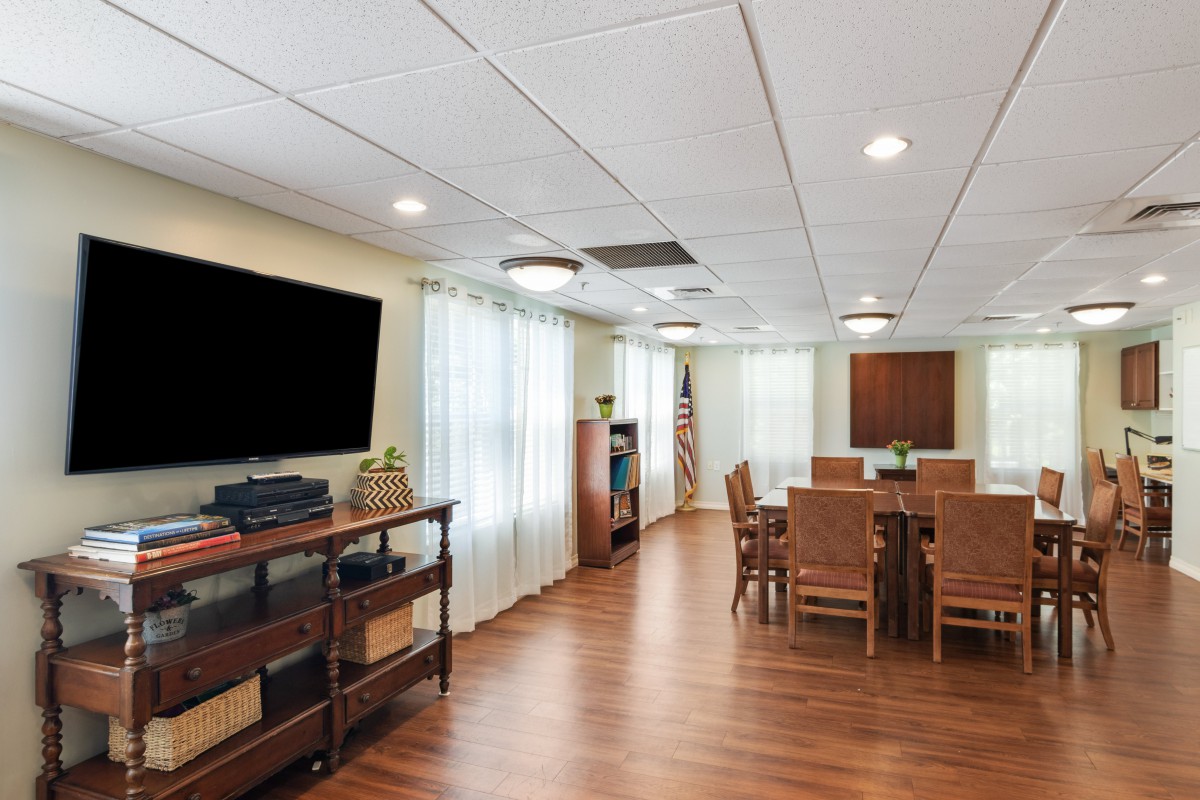 A well-lit conference room with wooden flooring, featuring a large television, a table surrounded by chairs, and decorative elements.