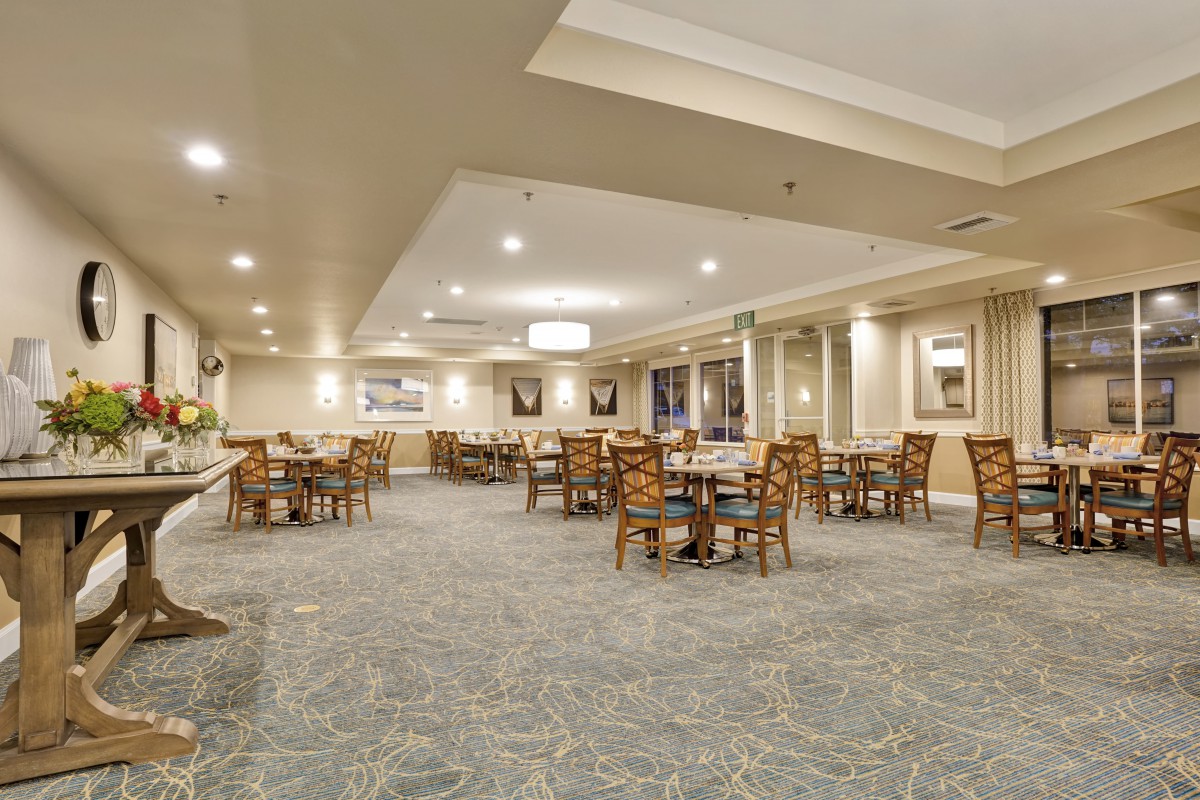 A spacious dining area with multiple tables and chairs, well-lit and featuring a carpeted floor.