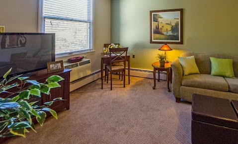 A cozy living room featuring a sofa, table with chairs, lamp, artwork on the wall, and a potted plant by the window.