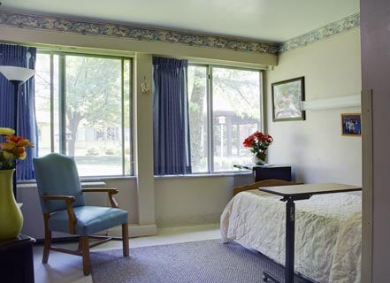 A hospital room with a bed, chair, and window providing natural light.