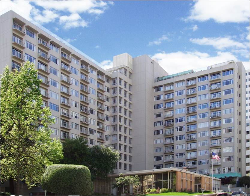 A tall apartment building with numerous windows, set against a bright blue sky and surrounded by greenery.